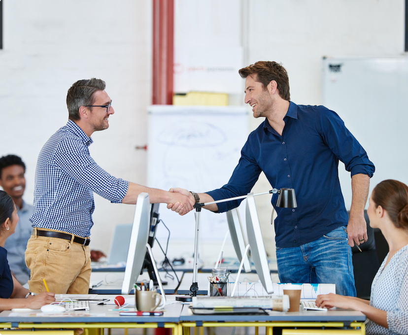 Two professionals shaking hands in a modern office setting, with colleagues seated at desks in the background.
